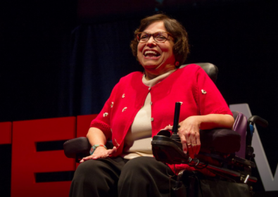 Judy Heumann sitting in her wheelchair on stage at TED with the words overlayed, "Remembering Judy Heumann 1947-2023."