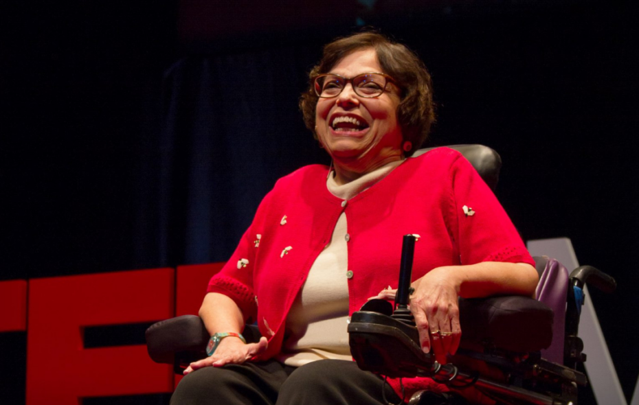 Judy Heumann sitting in her wheelchair on stage at TED with the words overlayed, "Remembering Judy Heumann 1947-2023."