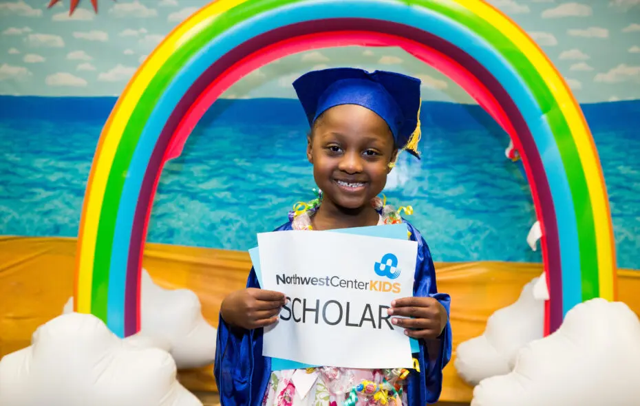Kid smiling wearing a graduation gown and holding a paper that reads: Northwest Center Kids Scholar.