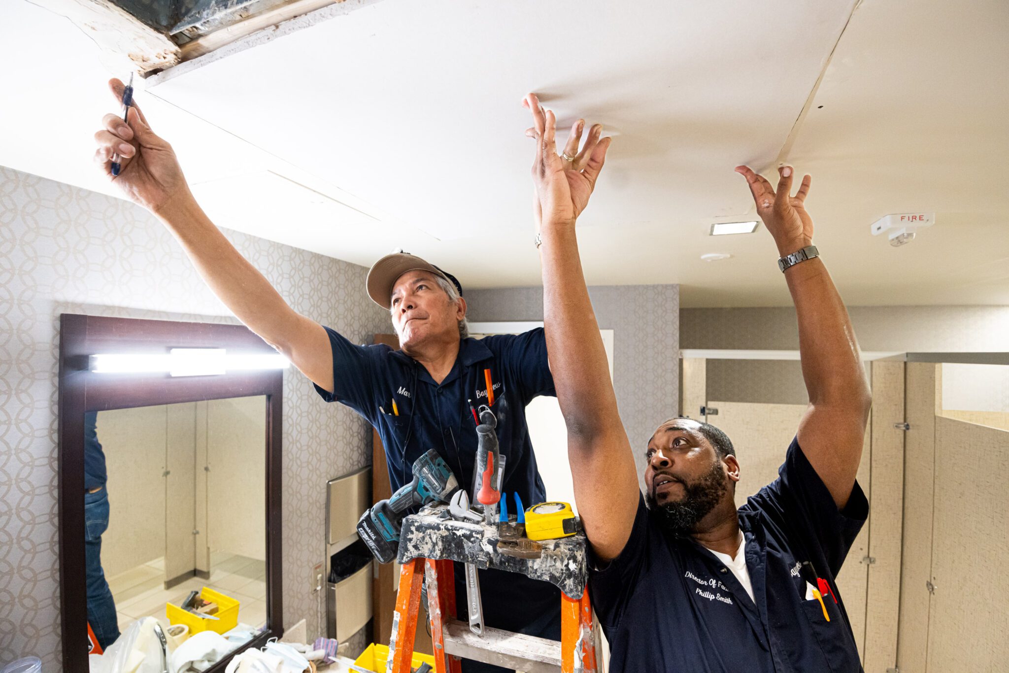 Two men standing on ladders and reaching up to repair a ceiling.