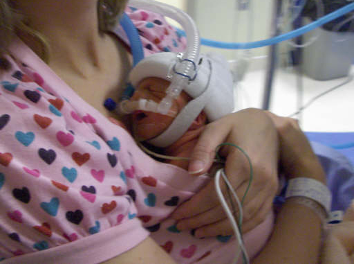 A close-up image in a hospital room shows a the shoulder of a white woman who is cradling a very small newborn baby to her chest. The newborn is wearing a protective cap and has breathing tubes coming out of his face. The woman wears a pink shirt covered in a heart pattern.